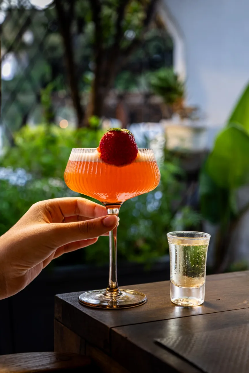 Cocktail à la fraise tenu à la main près d’un petit verre sur le comptoir en bois du Bar, devant la végétation